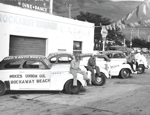 A black and white photo of men standing next to parked cars.